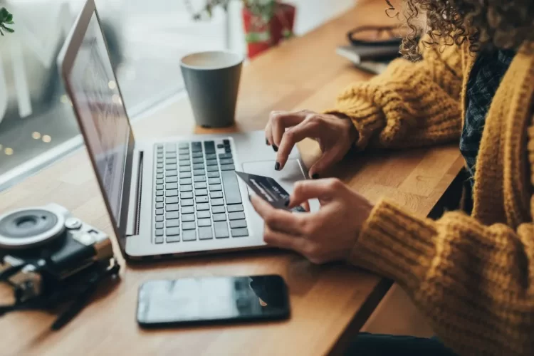 A imagem mostra uma pessoa, possivelmente amante de fotografia, porque há uma câmera do lado do notebook, comprando online. Ela está digitando os dados de seu cartão de crédito após se assegurar de que o site é realmente confiável para compras. 