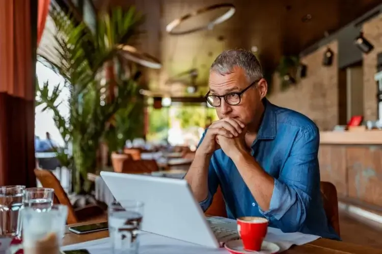 A imagem mostra um homem sentado diante de seu notebook em um café. Ele possivelmente está visitando um site, que foi mal otimizado e oferece uma experiência de navegação ruim. 