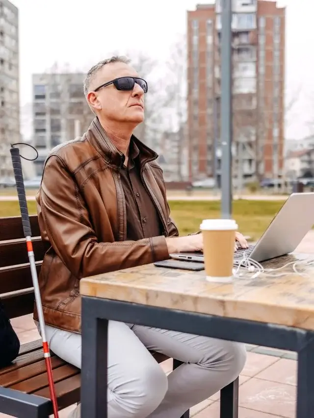 Um homem branco, de meia idade e com deficiência visual, está usando um notebook sobre a mesa de um parque. Ele está sentado em um banco e ao seu lado está uma bengala. Ao lado do notebook há um copo de café.