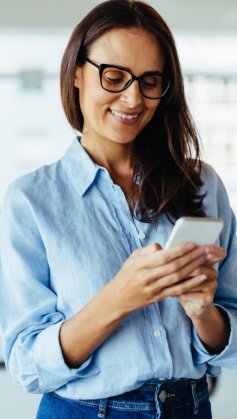 A imagem mostra uma mulher em um escritório mexendo em um smartphone. Ela possivelmente está gerenciando o perfil da empresa e aplicando algumas estratégias para conseguir seguidores no Instagram.