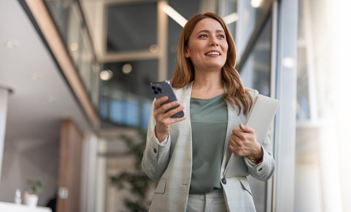 A imagem mostra uma mulher com roupa social no que parece ser o saguão de um escritório. Ela segura o celular em uma mão e uma pasta na outra, olhando para o horizonte enquanto pensa em formas de como monetizar no Instagram.