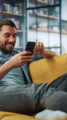 A imagem mostra um homem sorrindo enquanto toca a tela de seu smartphone. Ele possivelmente está em uma rede social e foi impactado pela publicação de uma empresa, que sabe quais são os melhores horários para postar no TikTok.