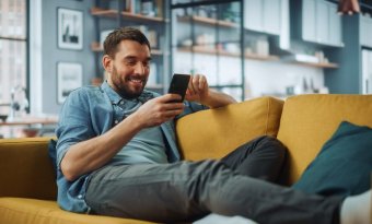 A imagem mostra um homem sorrindo enquanto toca a tela de seu smartphone. Ele possivelmente está em uma rede social e foi impactado pela publicação de uma empresa, que sabe quais são os melhores horários para postar no TikTok.