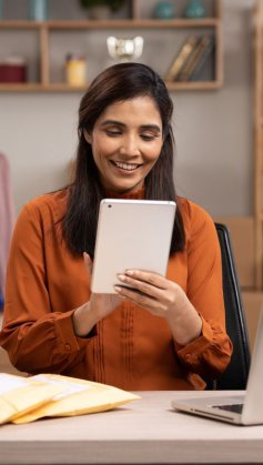 A imagem mostra uma mulher trabalhando em uma loja de roupas que aparentemente é virtual. Ela está segurando um tablet. Há uma mesa com pacotes e um notebook à frente dela, além de uma estante com caixas e uma arara com roupas penduradas.