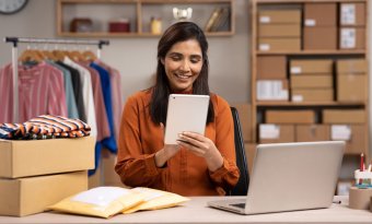 A imagem mostra uma mulher trabalhando em uma loja de roupas que aparentemente é virtual. Ela está segurando um tablet. Há uma mesa com pacotes e um notebook à frente dela, além de uma estante com caixas e uma arara com roupas penduradas.