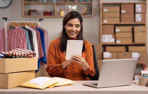 A imagem mostra uma mulher trabalhando em uma loja de roupas que aparentemente é virtual. Ela está segurando um tablet. Há uma mesa com pacotes e um notebook à frente dela, além de uma estante com caixas e uma arara com roupas penduradas.