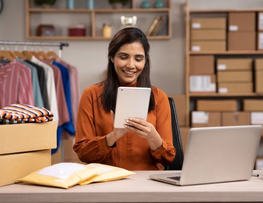 A imagem mostra uma mulher trabalhando em uma loja de roupas que aparentemente é virtual. Ela está segurando um tablet. Há uma mesa com pacotes e um notebook à frente dela, além de uma estante com caixas e uma arara com roupas penduradas.