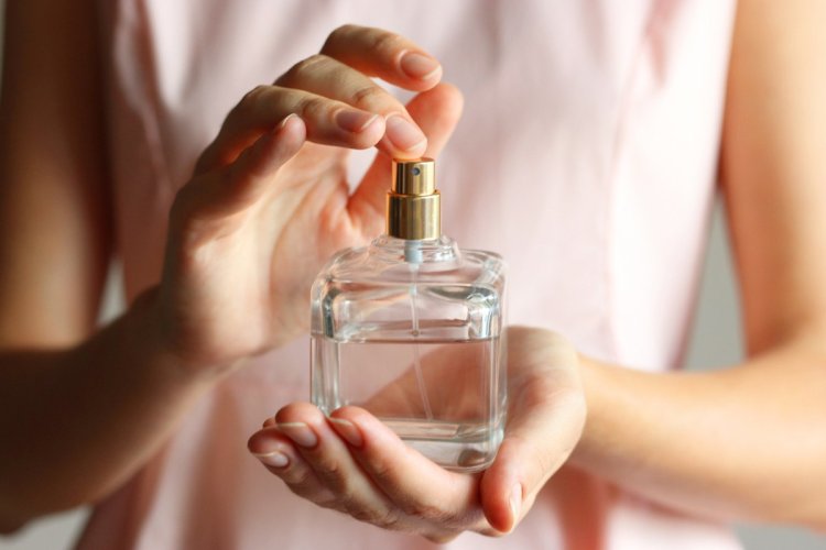 A imagem mostra uma mulher segurando um frasco de perfume. O rosto dela não aparece. Ela está borrifando um pouco do produto no punho para sentir o aroma. 