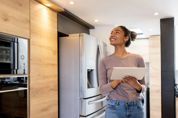 A imagem mostra uma mulher andando pela cozinha da casa dela. Ela está segurando um tablet, possivelmente para fazer as configurações dos dispositivos inteligentes que vão controlar alguns aspectos da residência. 