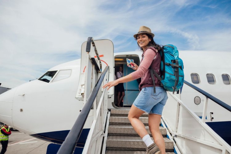 A imagem mostra uma mulher embarcando em um avião. Ela usa um chapéu e uma mochila azul, que são acessórios bastante buscados por quem ama viajar. 