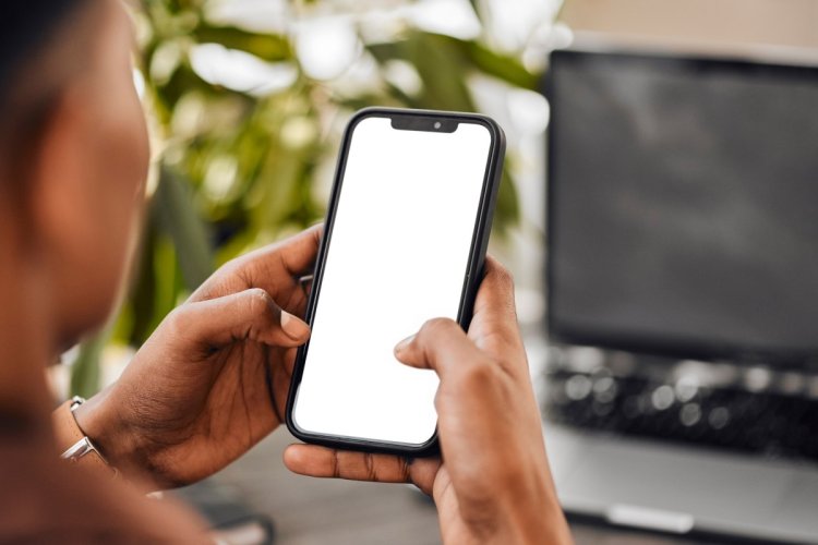 A imagem mostra um homem com um celular na mão. O rosto dele não aparece. Ele possivelmente está usando o aparelho no ambiente de trabalho, já que há um notebook desfocado no fundo. 