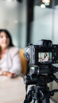 A imagem mostra uma mulher gravando um vídeo com uma câmera. Ela aparece desfocada, já que a foto está mais concentrada na câmera. Ela parece estar gravando um VSL.