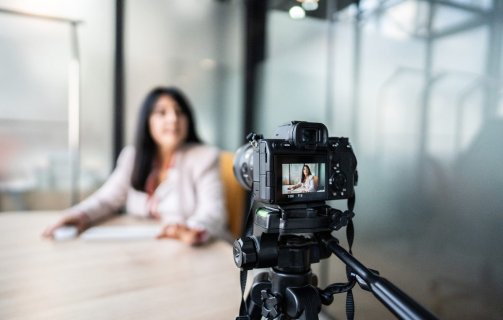 A imagem mostra uma mulher gravando um vídeo com uma câmera. Ela aparece desfocada, já que a foto está mais concentrada na câmera. Ela parece estar gravando um VSL.