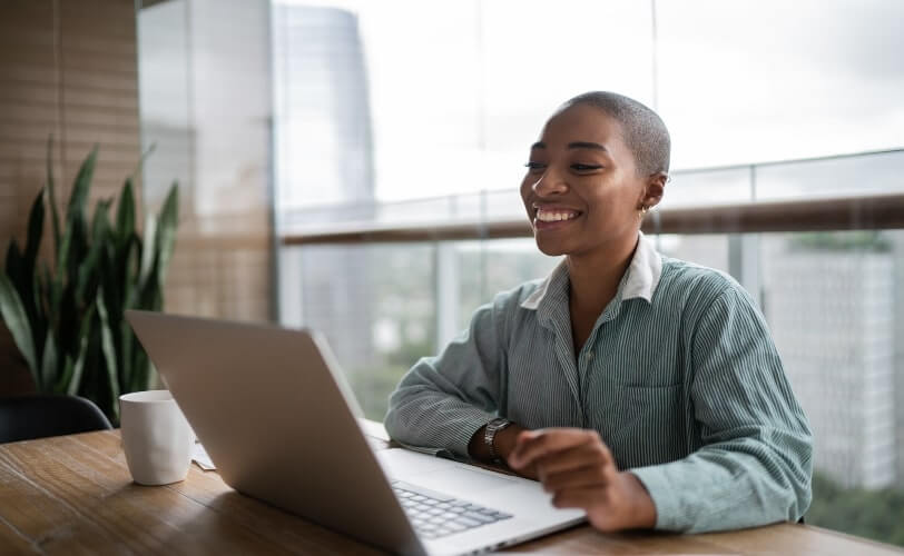 Imagem de uma mulher afro-americana sentada à mesa com um notebook aberto e uma xícara ao lado. A mulher sorri para a tela do computador.