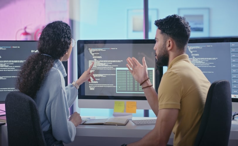 Dois jovens programadores, sendo um homem branco, de costas, e uma mulher afro-americana, de lado; ambos vestindo roupas casuais, estão de frente para monitores observando linhas de código e usando Flask Python.