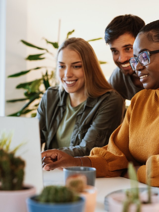 Um trio multiétnico de pessoas, sendo duas mulheres e um homem, está em frente a um notebook. Eles trabalham em um escritório de empresa, vestindo roupas casuais e sorrindo.