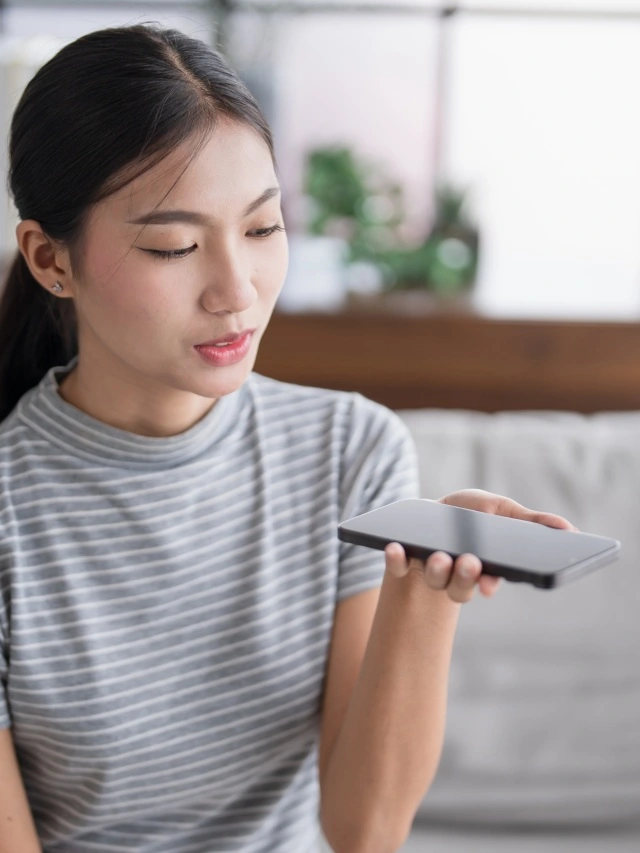 Uma jovem mulher asiática, vestindo camiseta listrada em cinza e branco, segura um smartphone, no qual usa um assistente de IA para conversar.