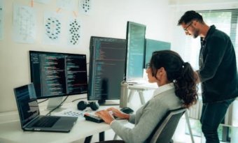 A imagem mostra um homem e uma mulher conversando na frente de um computador com várias telas. Há alguns códigos nelas, e eles possivelmente estão debatendo sobre formas de aplicar o Machine Learning na empresa onde trabalham.