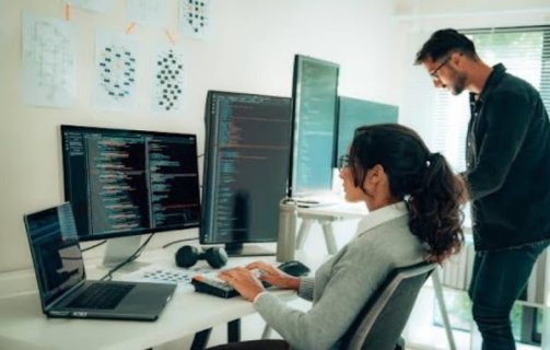 A imagem mostra um homem e uma mulher conversando na frente de um computador com várias telas. Há alguns códigos nelas, e eles possivelmente estão debatendo sobre formas de aplicar o Machine Learning na empresa onde trabalham.