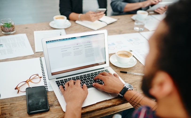 Um homem está acessando uma página na internet em seu notebook, posicionado em uma mesa com uma xícara de café, óculos, smartphone e papeis ao redor. A página virtual acessada por ele possui em sua URL um favicon.