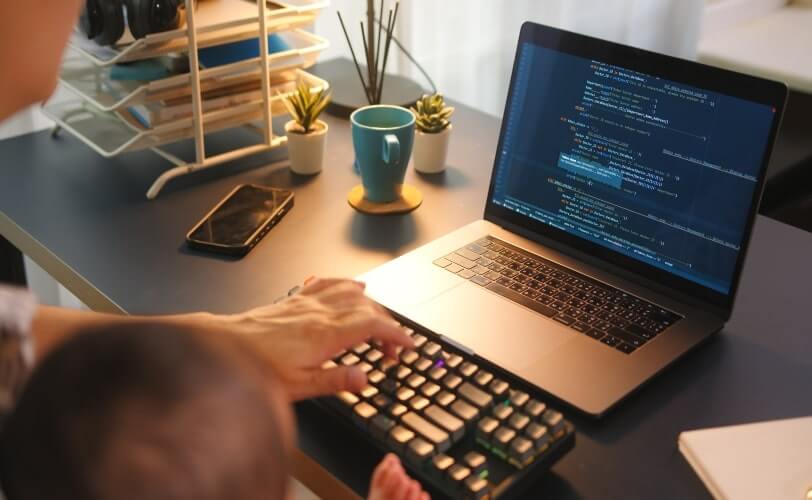 A imagem mostra uma pessoa programando em um laptop enquanto cuida de um bebê. O ambiente de trabalho inclui uma mesa organizada com um teclado externo, uma xícara de café, plantas decorativas e um smartphone ao lado. O código na tela indica um ambiente de desenvolvimento de software, usando uma das várias bibliotecas Python. 