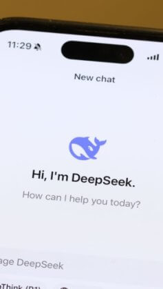 Imagem mostra um smartphone exibindo o logotipo do "DeepSeek", sendo segurado por uma mão de uma pessoa branca. O design sugere uma identidade visual moderna e tecnológica, relacionada a uma ferramenta de inteligência artificial.