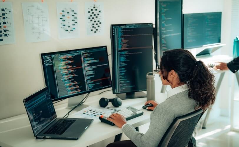 Uma jovem mulher de perfil, vestindo suéter cinza e camisa branca, está em frente a dois monitores de computador e um notebook. Ela digita linhas de código e avalia um a um, usando uma linguagem de programação.